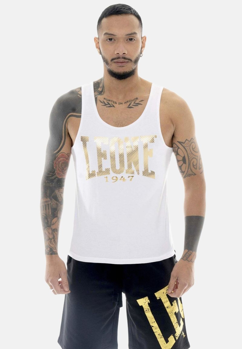 Homme avec des tatouages porte un débardeur blanc avec le texte doré "LEONE 1947" et un short noir avec une grande impression dorée "LEO" sur le côté.