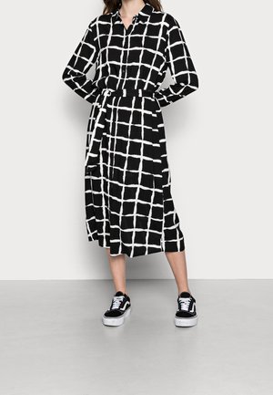 Femme portant une robe chemise midi noire à motif quadrillé blanc et des baskets noires, debout les mains sur les hanches devant un fond uni.