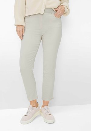 Billedet viser personens ben iført lysegrå skræddersyede bukser, beige sneakers og en cremefarvet sweater, stående med den ene hånd i lommen.