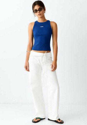 Mujer con gafas de sol oscuras, top azul sin mangas, pantalones anchos blancos con cordón y chanclas negras, de pie sobre fondo liso.