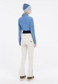 Hellblau gestreiftes Langarmoberteil, weiße Hose mit Reißverschlüssen an den Knöcheln und dunkelgrüne Schuhe. Die Mütze hat blau- und grautöne.