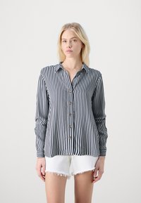Camisa de botones a rayas en azul marino y blanco con cuello, mangas largas y cinco botones en la parte delantera. Llevada con pantalones cortos blancos deshilachados.