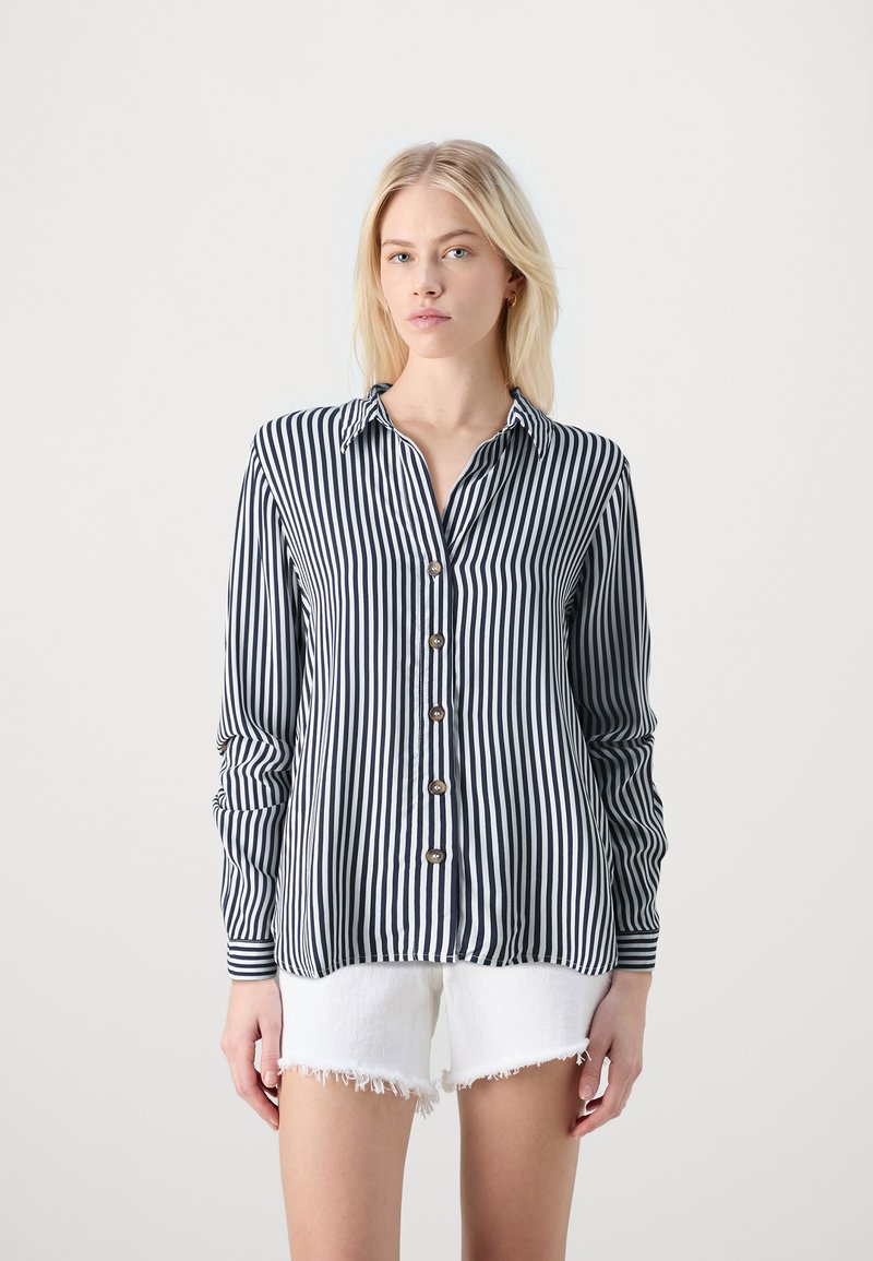 Camisa de botones a rayas en azul marino y blanco con cuello, mangas largas y cinco botones en la parte delantera. Llevada con pantalones cortos blancos deshilachados.