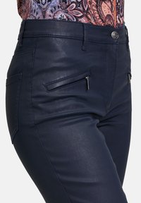 Marineblaue, high-waisted Hose aus glattem, glänzendem Material mit zwei vorderen Reißverschlusstaschen und einem einzigen Knopfverschluss.