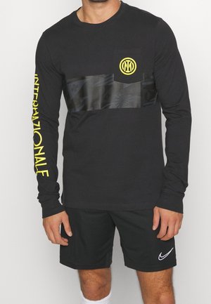 Homme portant un t-shirt noir à manches longues avec le texte jaune "Internazionale" sur la manche et un logo sur la poitrine, associé à un short noir Nike.