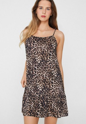 Frau mit langen Haaren, die ein ärmelloses Kleid mit Leopardenmuster und dünnen Trägern trägt, steht vor einem einfarbigen hellen Hintergrund.