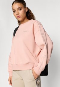 Rózsaszín kapucnis pulóver laza szabással, logó hímzéssel a mellkason, díszítő oldalcsíkokkal. Bézs nadrággal és fekete hátizsákkal viselve.