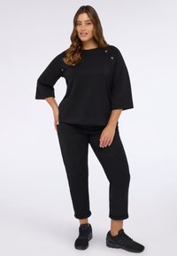Czarny longsleeve z otworami, okrągłym dekoltem i luźnym fasonem, w połączeniu z czarnymi spodniami 3/4 i czarnymi trampkami.