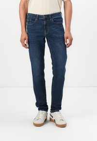 Jeans de mezclilla azul oscuro con corte ajustado, que cuentan con cinco bolsillos, cremallera y detalles de costura sutiles. Llevados con zapatillas blancas.