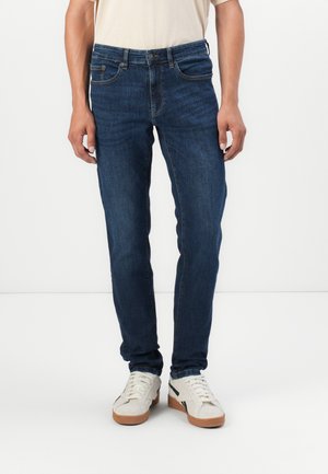 Jeans de mezclilla azul oscuro con corte ajustado, que cuentan con cinco bolsillos, cremallera y detalles de costura sutiles. Llevados con zapatillas blancas.
