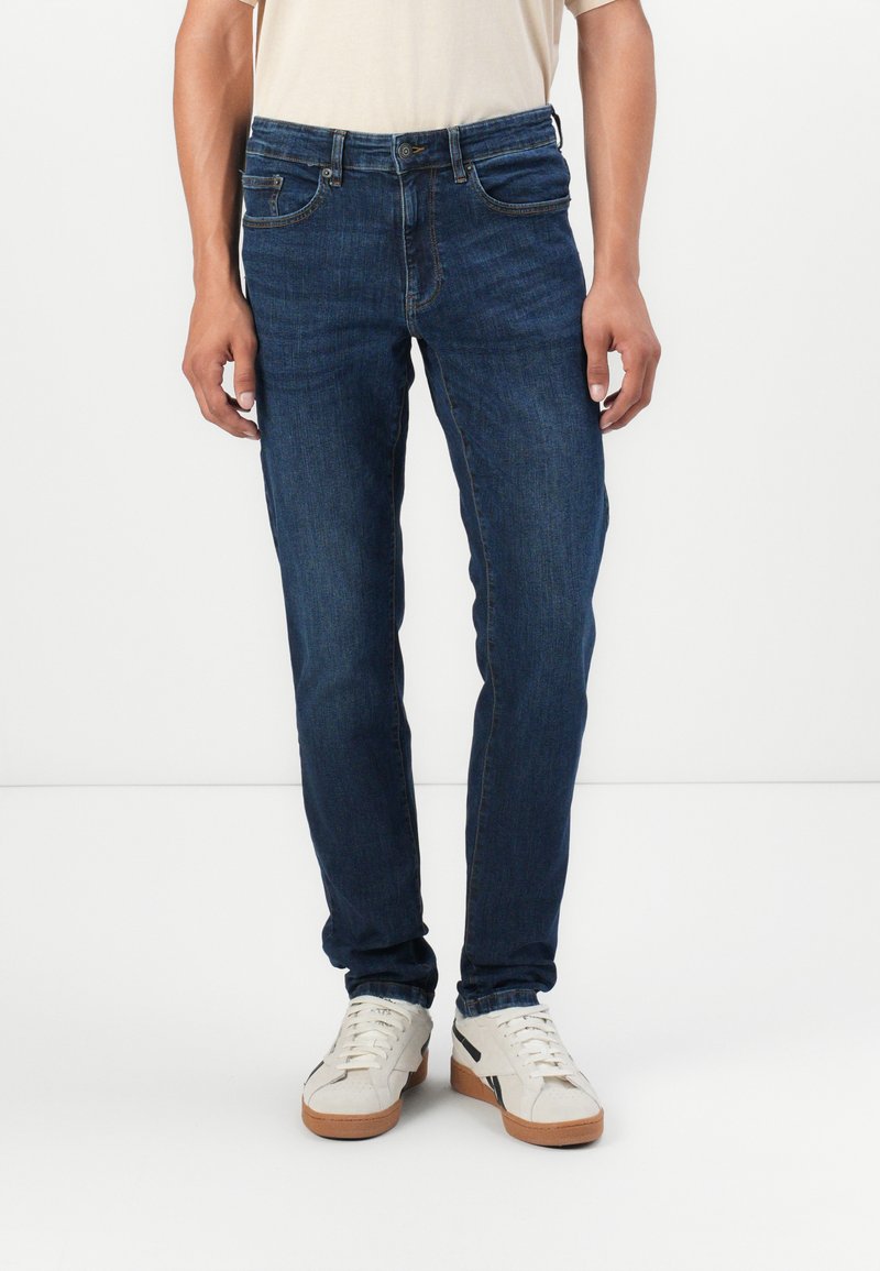 Jeans de mezclilla azul oscuro con corte ajustado, que cuentan con cinco bolsillos, cremallera y detalles de costura sutiles. Llevados con zapatillas blancas.
