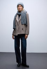 Abrigo de lana gris claro y texturizado con doble botonadura, combinado con una bufanda y un gorro a juego; vaqueros de pierna ancha en azul oscuro y botas negras.