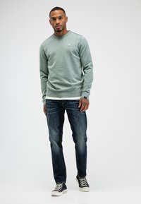 Grüner Strickpullover mit Rundhalsausschnitt, ribbelten Bündchen und Saum; kombiniert mit dunkelblauen Jeans und schwarzen Sneakers mit weißen Sohlen.