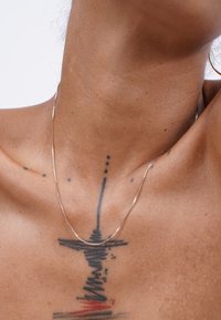 Dünne, zarte Halskette aus Gold, die auf der Haut mit einem Tattoo ruht. Sie hat eine glatte, glänzende Oberfläche und ein dezentes Design.