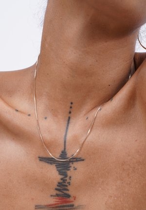 Thin, delicate gold chain necklace resting on skin with a tattoo. Features a smooth, shiny surface and subtle design.