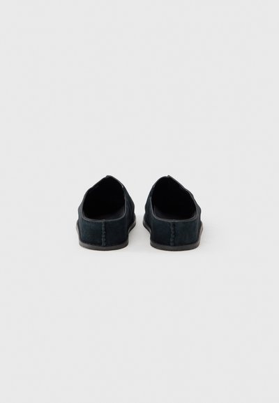 Chaussures en daim noir sans lacets avec une silhouette basse, une texture douce et des surpiqûres apparentes le long des coutures arrière et latérales.