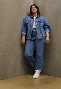 Veste en denim sur t-shirt blanc, associée à un jean en denim taille haute. Baskets blanches, se tenant contre un mur gris texturé.