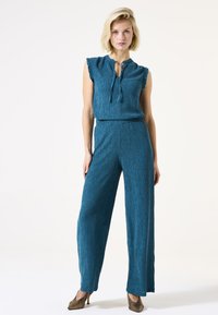 Teal, gestructureerde jumpsuit met een ruchesdecolleté en een strikdetail. Wijde broekspijpen met een losse pasvorm. Draag met bronzen, gestructureerde hakken.