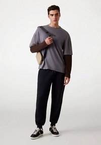 Camicia grigia a maniche corte indossata sopra una camicia marrone a maniche lunghe, abbinata a pantaloni sportivi neri e sneakers nere. Indossata una piccola borsa a tracolla beige.