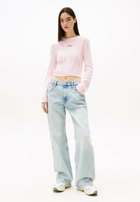 Suéter corto de punto trenzado en rosa claro con un pequeño logo rojo, combinado con jeans de pierna ancha en azul claro y zapatillas blancas.