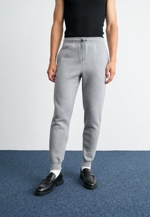 Personne portant un jogging gris avec cordon, un haut noir sans manches, des chaussettes blanches et des mocassins noirs à enfiler, debout sur un tapis bleu.