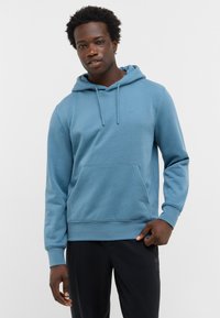 Mężczyzna w jasnoniebieskiej bluzie i czarnych spodniach stoi na tle jednolitego, jasnego tła, zwrócony przodem z odprężonym wyrazem twarzy.