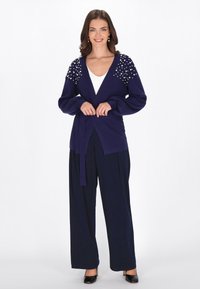 Cardigan marine avec un décolleté en V profond, orné de détails en perles aux épaules, noué à la taille, associé à un pantalon évasé et des talons.