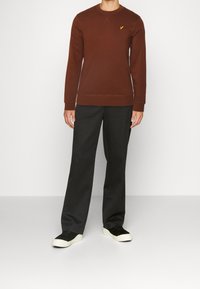 Sudadera marrón con cuello redondo y detalles de costura, combinada con pantalones gris oscuro y zapatillas negras con suela blanca.