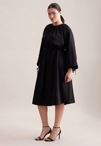 Zwarte midijurk met lange mouwen en een geplooide halslijn. Heeft een strik in de taille en een gladde stof. Gekleed met zwarte sandaaltjes met hoge hakken.