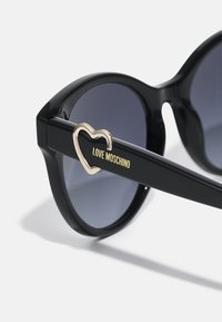 Fekete, túlméretezett napszemüveg szürke lencsékkel, melynek fekete karján egy szív alakú, arany kiegészítő található, amelyre az "LOVE MOSCHINO" felirat van gravírozva.
