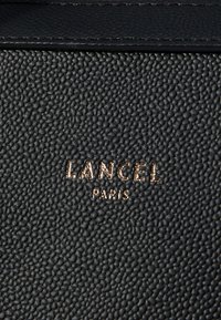 Superficie de cuero negro texturizado con un logo dorado estampado que dice "LANCEL PARIS" en el centro inferior.