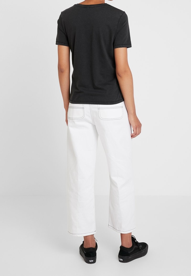 Personne debout, de dos, portant un t-shirt noir à manches courtes, un pantalon blanc à jambes larges avec des poches arrière, et des baskets noires.
