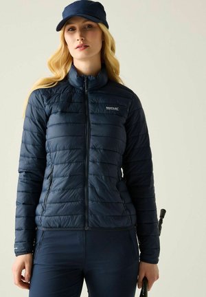 Vrouw met een marineblauw donsjack en bijpassend petje, met een wandelstok tegen een effen lichtgrijze achtergrond.