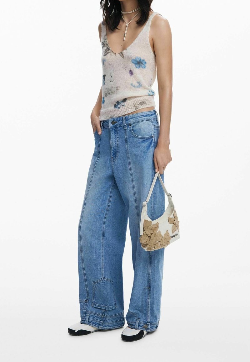 Femme portant un top sans manches fleuri, un jean large bleu, des chaussures blanches et noires, tenant un petit sac à main blanc avec des détails de fleurs beige.
