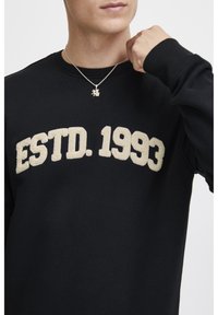Crna dukserica s debelim, teksturiranim, kremastim printom "ESTD. 1993" preko prednje strane. Ima okrugli izrez i rebraste rukave.