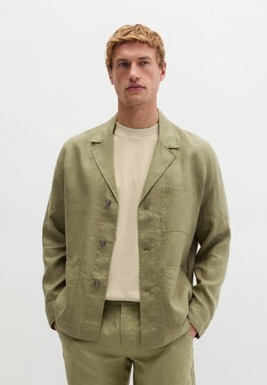 Junger Mann trägt einen olivgrünen Leinenanzug mit aufgesetzten Taschen und einem beigen T-Shirt mit Rundhalsausschnitt, steht mit den Händen in den Taschen.