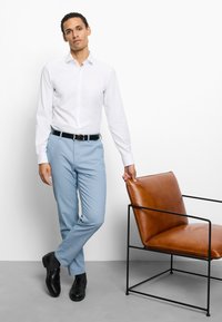 Homme en chemise blanche et pantalon bleu clair debout à côté d'une chaise en cuir marron avec une armature métallique noire.