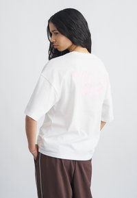 Νέα γυναίκα με μακριά σκουρόχρωμα μαλλιά, φορώντας ένα άνετο λευκό μπλουζάκι και καφέ παντελόνια, που στέκεται μπροστά σε ουδέτερο ανοιχτό φόντο.