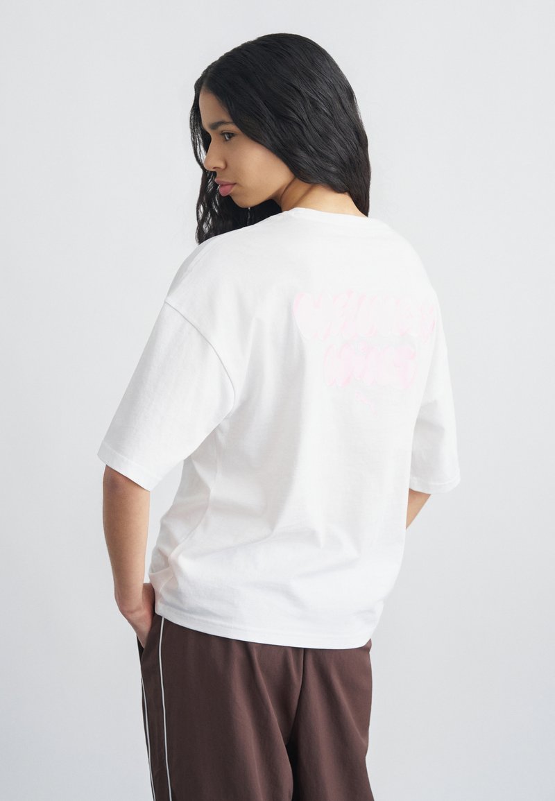 Νέα γυναίκα με μακριά σκουρόχρωμα μαλλιά, φορώντας ένα άνετο λευκό μπλουζάκι και καφέ παντελόνια, που στέκεται μπροστά σε ουδέτερο ανοιχτό φόντο.