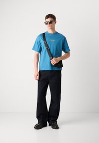 Teal színű pamut póló Calvin Klein logóval, sötét, bő szárú nadrággal és fekete bőrcipővel párosítva, feketét válltáskával kiegészítve.