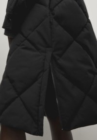Zwarte gewatteerde pufferjas met een ritssluiting aan de achterkant en diagonale stikpatronen. Het materiaal is zacht met een gladde textuur.