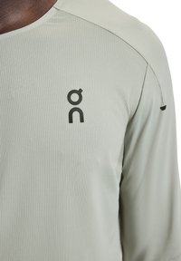 Camicia a maniche lunghe grigio chiaro con tessuto testurizzato e logo nero. Presenta cuciture piatte e scollo rotondo, enfatizzando la funzionalità.