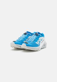 Une paire de chaussures de trail Salomon bleues et blanches avec un système de lacets rapide et des semelles texturées sur fond blanc.