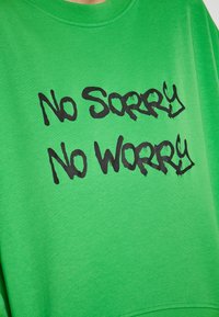 Sudadera verde brillante de algodón con un corte relajado. Presenta un llamativo texto en negro estilo graffiti: "No disculpas, sin preocupaciones" impreso de manera prominente.