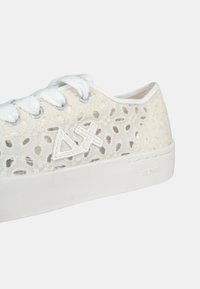Zapatillas de cordones en beige claro con un patrón perforado, suela de goma blanca y detalle de logo bordado. Presenta cordones y ojetes blancos.