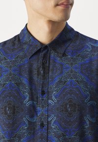 Koszula z krótkim rękawem w czarnym materiale z niebieskim i zielonym, skomplikowanym wzorem paisley, z zapiętym na guziki kołnierzem i plisą z przodu.