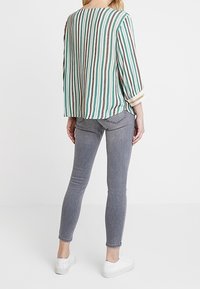 Blusa a righe in teal, bianco e marrone con maniche a tre quarti; abbinata a jeans skinny grigi e sneakers bianche. Sfondo semplice.