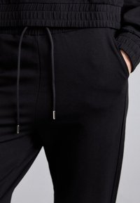 Asmuo vilkintis juodus treniruočių kelnes su raišteliu, ranka įsidėjusi į kišenę, o atitinkama juoda elastinga liemenė dalinai matoma.