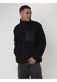 Uomo che indossa una giacca in pile nera con zip e tasca sul petto, che guarda di lato con le mani vicino alle tasche dei jeans.
