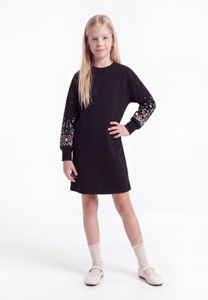 Mädchen mit langen blonden Haaren trägt ein schwarzes Kleid mit bunter Blumenstickerei an den Ärmeln, cremefarbene Socken und cremefarbene Schuhe, steht mit der Hand in der Hüfte.